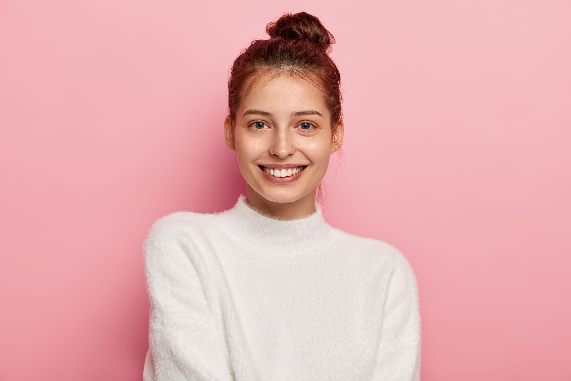 Beauty and facial expressions concept. Tender feminine woman with blue eyes, smiles pleasantly, has toothy smile, wears white comfortable sweater, looks directly at camera, isolated on pink background
